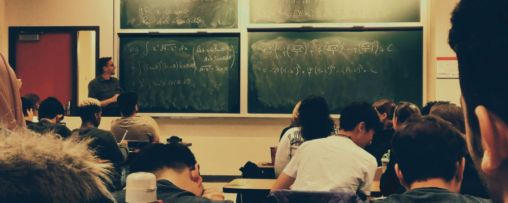 Students sitting at desks with a techer writting on chalkboard